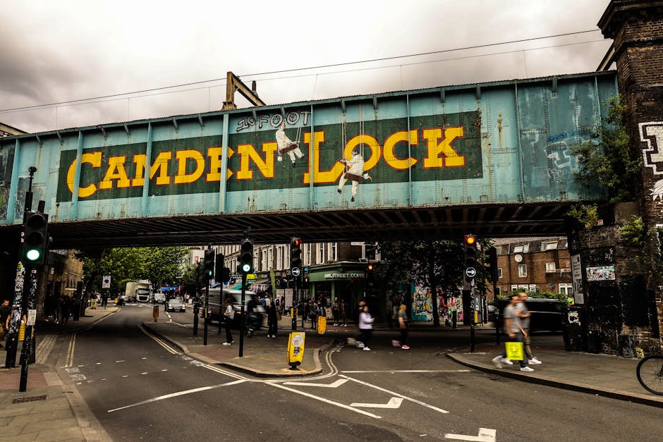 A large, horizontally oriented metal railway bridge painted in faded turquoise with rust and weathering visible on its surface, spans across a busy street scene. The bridge features bold yellow lettering spelling 'CAMDEN LOCK' and has two painted illustrations of children hanging from the letters. Above the sign, a small footprint graphic is visible with the words '2 FOOT' written nearby. Below the bridge, there is a street intersection with multiple traffic lights, pedestrian crossings, and a variety of pedestrians walking in different directions. Some of the pedestrians are blurred due to motion, and the scene includes typical urban elements such as bicycles, parked vehicles, and shopfronts in the background. On the right side, part of a brick building and some green foliage are visible, contributing to the cityscape environment. This image relates to urban waste or rubbish disposal contexts, as Waste Disposal Belsize Park operates in such environments, often involving on-site clearance or private waste handling in city streets.