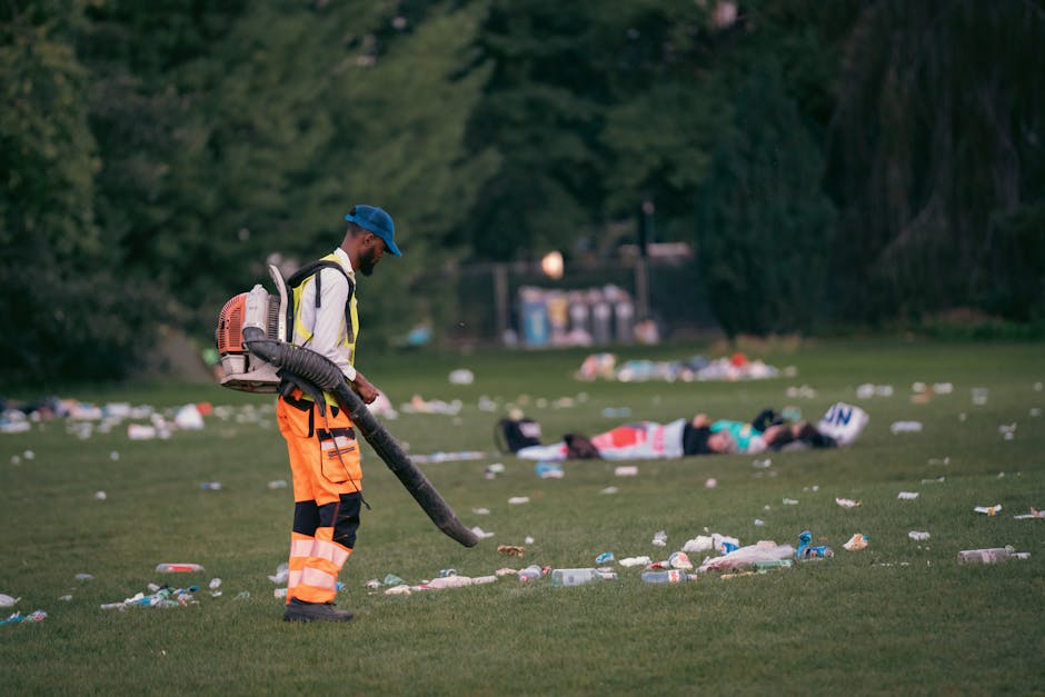 A municipal worker wearing a high-visibility orange and black uniform with a blue cap operates a leaf blower or vacuum device to clear litter from a grassy park area. The worker is positioned in the foreground, standing on a patch of green grass, with a backpack-style vacuum or blower hose directed downward to pick up scattered trash. In the background, blurred figures of two individuals lie on the grass amid additional debris, with several plastic bottles, paper waste, and other refuse spread across the lawn. Tall trees with dark foliage border the scene, and beyond them, a small structure with containers or bins is visible under the late afternoon or early evening light, creating a calm but cluttered environment. The image illustrates private waste collection or on-site rubbish clearance in a public outdoor space, emphasizing the importance of proper rubbish management by waste disposal services such as Waste Disposal Belsize Park.