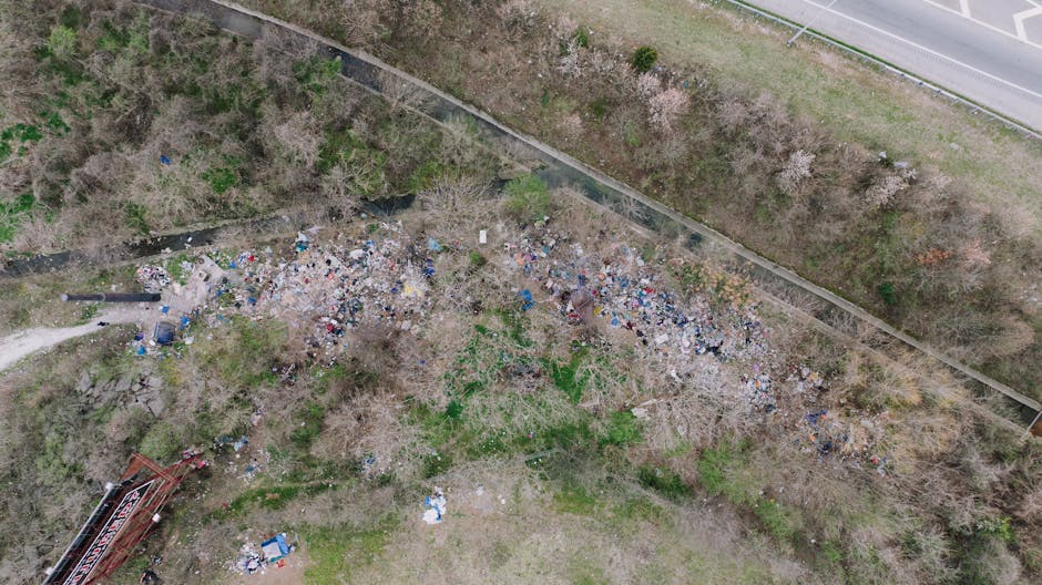 An aerial view shows a large, irregularly shaped accumulation of mixed waste and rubbish scattered across a patch of land, with various materials including plastics, paper, and other debris visible. The waste appears loosely piled, with some items overlapping and spreading out in different directions. Surrounding the rubbish are leafless trees and sparse greenery, indicating a late autumn or winter season. To the upper right, a paved road runs parallel to a chain-link fence that borders the area, separating it from the roadside. In the lower left corner, a small portion of industrial equipment or a waste container is partially visible. The scene is lit with natural daylight, highlighting the contrasting textures of the trash against the bare branches, and reflects an informal or unmanaged waste accumulation site, which Waste Disposal Belsize Park may handle through private collection or on-site clearance services as part of its rubbish removal solutions.