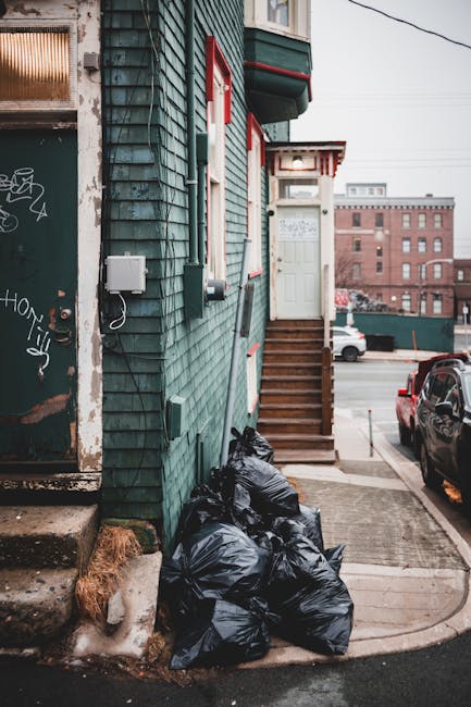 The image depicts a small urban building with a teal-colored wooden exterior, featuring shingle siding and a white door accessed by a set of wooden stairs. Several black garbage bags are stacked on the concrete pavement near the base of the stairs, indicating disposal or rubbish collection, possibly by an independent waste removal service. The building is situated on a paved street corner, with a parking area visible on the right side containing two parked cars, one black and one red. To the left, there is a graffiti-covered metal door with a small step leading up to it, alongside an electrical box mounted on the building's wall. In the background, there are several mid-rise brick buildings and overcast skies, conveying an urban environment. The scene emphasizes private waste handling within a residential or commercial setting, with the rubbish bags prepared for collection. Waste Disposal Belsize Park's services are supported by the context, highlighting the importance of efficient rubbish removal in city environments.