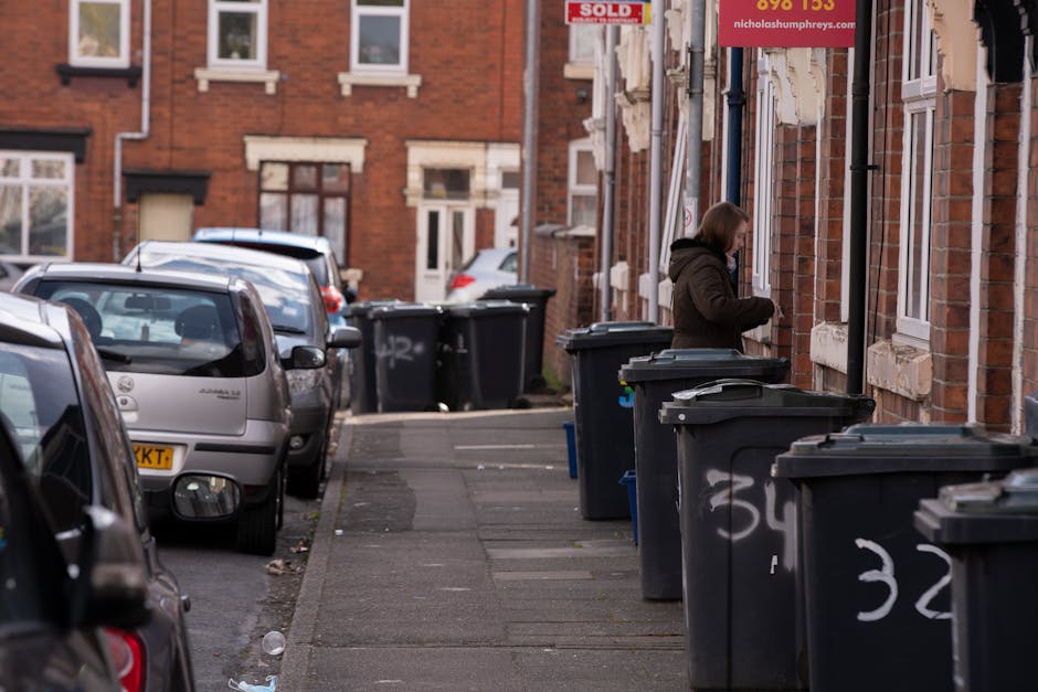 A woman standing on a concrete sidewalk next to a row of dark grey wheelie bins with closed lids, positioned along the exterior wall of a red brick residential building. The bins are marked with white spray-painted numbers and appear to be part of private waste collection service. The woman, dressed in a dark brown jacket, is loading or managing waste inside one of the bins. Parked cars are lined up along the curb on the street adjacent to the sidewalk, including a silver hatchback closest to the camera, with more vehicles further down. In the background, additional residential buildings with white-framed windows and small front yards can be seen, along with a partially visible 'sold' sign attached to the building. The scene is illuminated by natural daylight, with some shadows cast by the parked cars and the nearby buildings, illustrating an urban setting focused on alternative waste handling and rubbish disposal at the residential level.