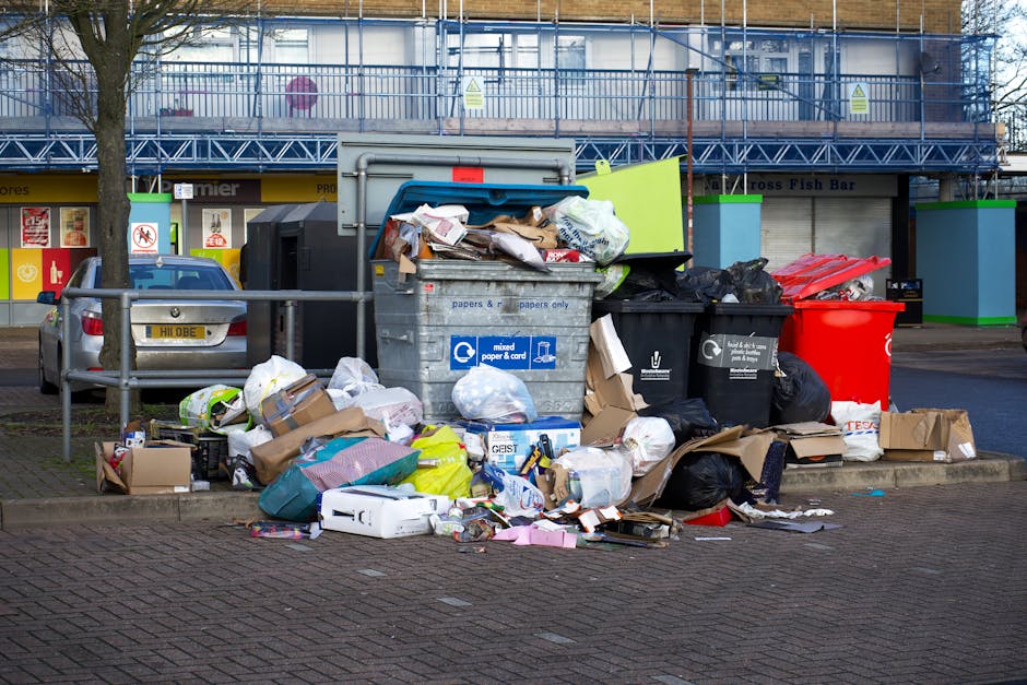 The image depicts a small urban building with a teal-colored wooden exterior, featuring shingle siding and a white door accessed by a set of wooden stairs. Several black garbage bags are stacked on the concrete pavement near the base of the stairs, indicating disposal or rubbish collection, possibly by an independent waste removal service. The building is situated on a paved street corner, with a parking area visible on the right side containing two parked cars, one black and one red. To the left, there is a graffiti-covered metal door with a small step leading up to it, alongside an electrical box mounted on the building's wall. In the background, there are several mid-rise brick buildings and overcast skies, conveying an urban environment. The scene emphasizes private waste handling within a residential or commercial setting, with the rubbish bags prepared for collection. Waste Disposal Belsize Park's services are supported by the context, highlighting the importance of efficient rubbish removal in city environments.